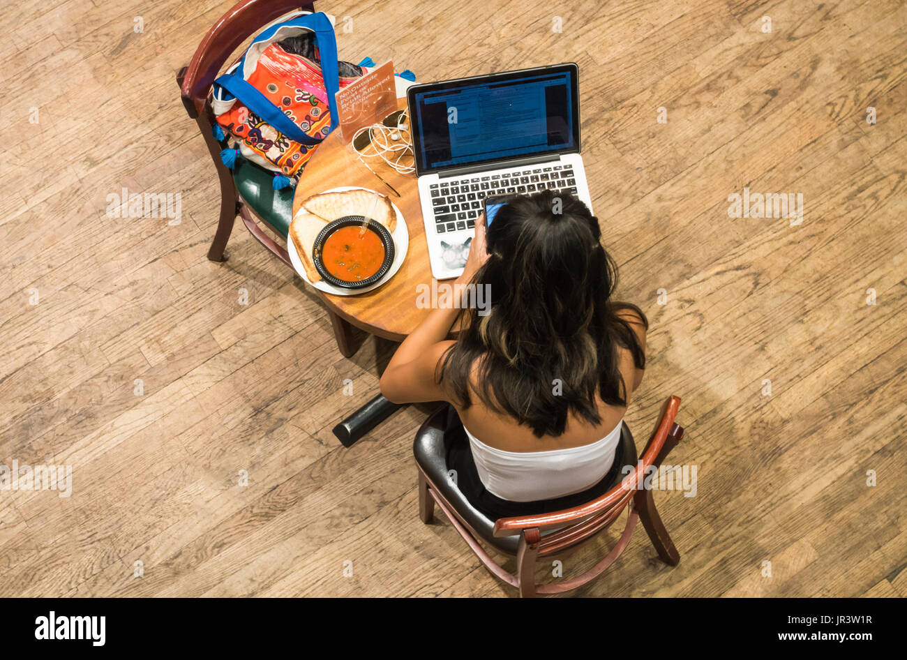 An over view of a young woman using her smartphone and laptop while having lunch at Housing Works Bookstore Cafe in SoHo in New York City Stock Photo