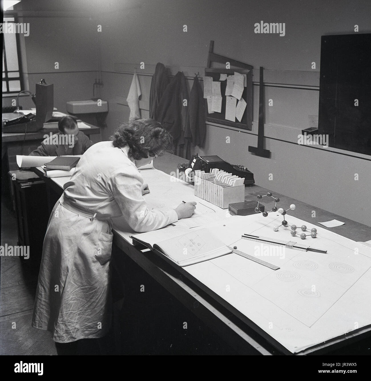1951, historical, female mathematician in white coat at a workbench doing calculations in a work book using, a ruler, a slide-rule and a ball and stick model, England, UK. Stock Photo