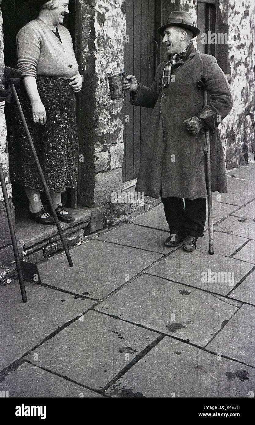 1940s, historical, elderly man with wearing an overcoat and hat and holding onto a walking stick, chats to a lady at the entrance to her cottage about her buying some of his cockles. Stock Photo