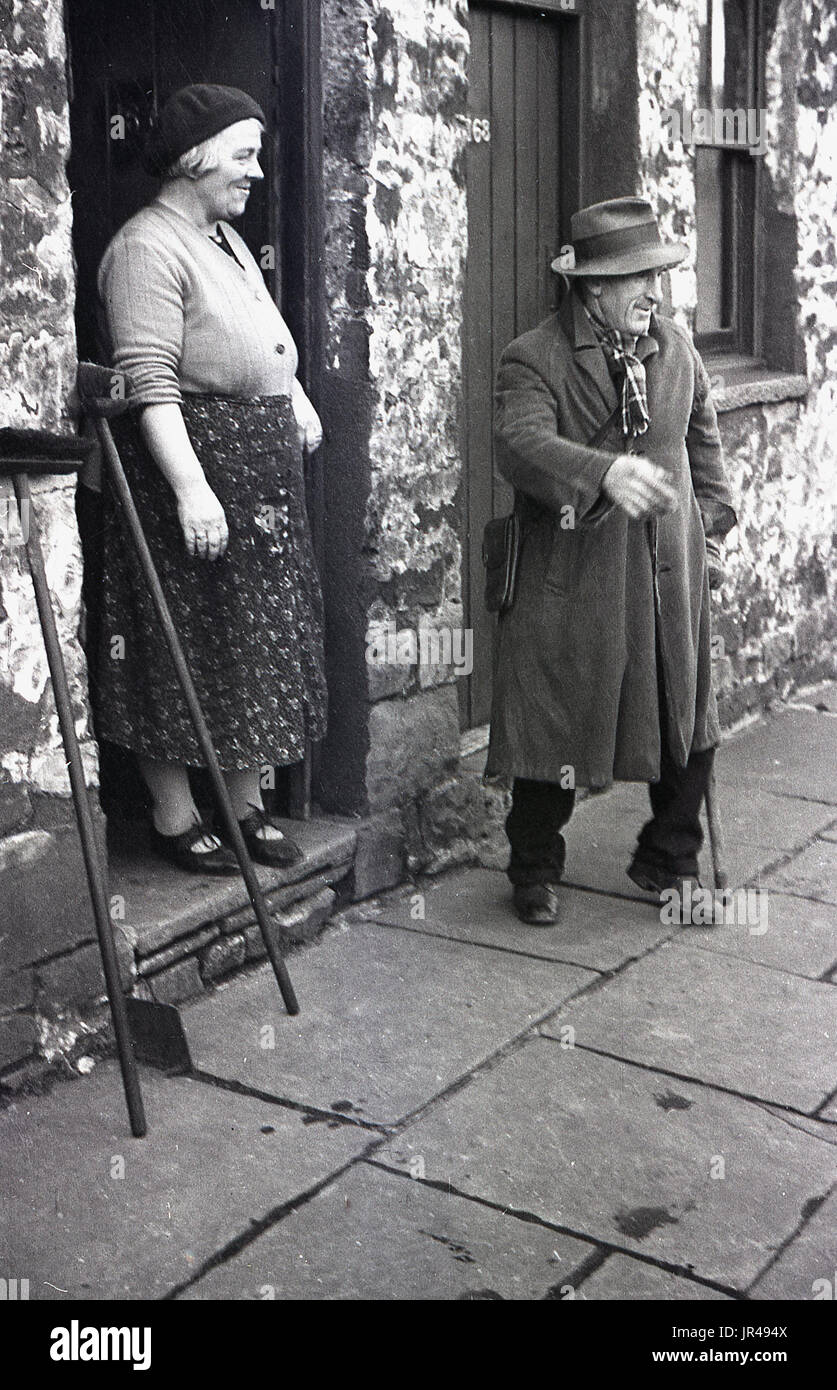 1940s, historical, elderly gentleman wearing an overcoat and hat and lady in beret standing at the entrance to her stone cottage, Merthyr Tydfil, South Wales, UK. The man is attempting to sell cockles he has sourced on the local beach door to door. Stock Photo