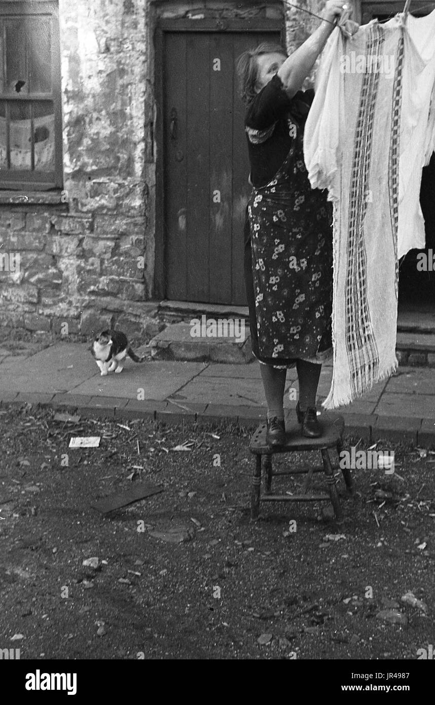 1940s, Merthyr Tydfil, South Wales, lady in apron standing on a wooden stool hanging up wet clothes to dry in the street outside her stone cottage. Stock Photo