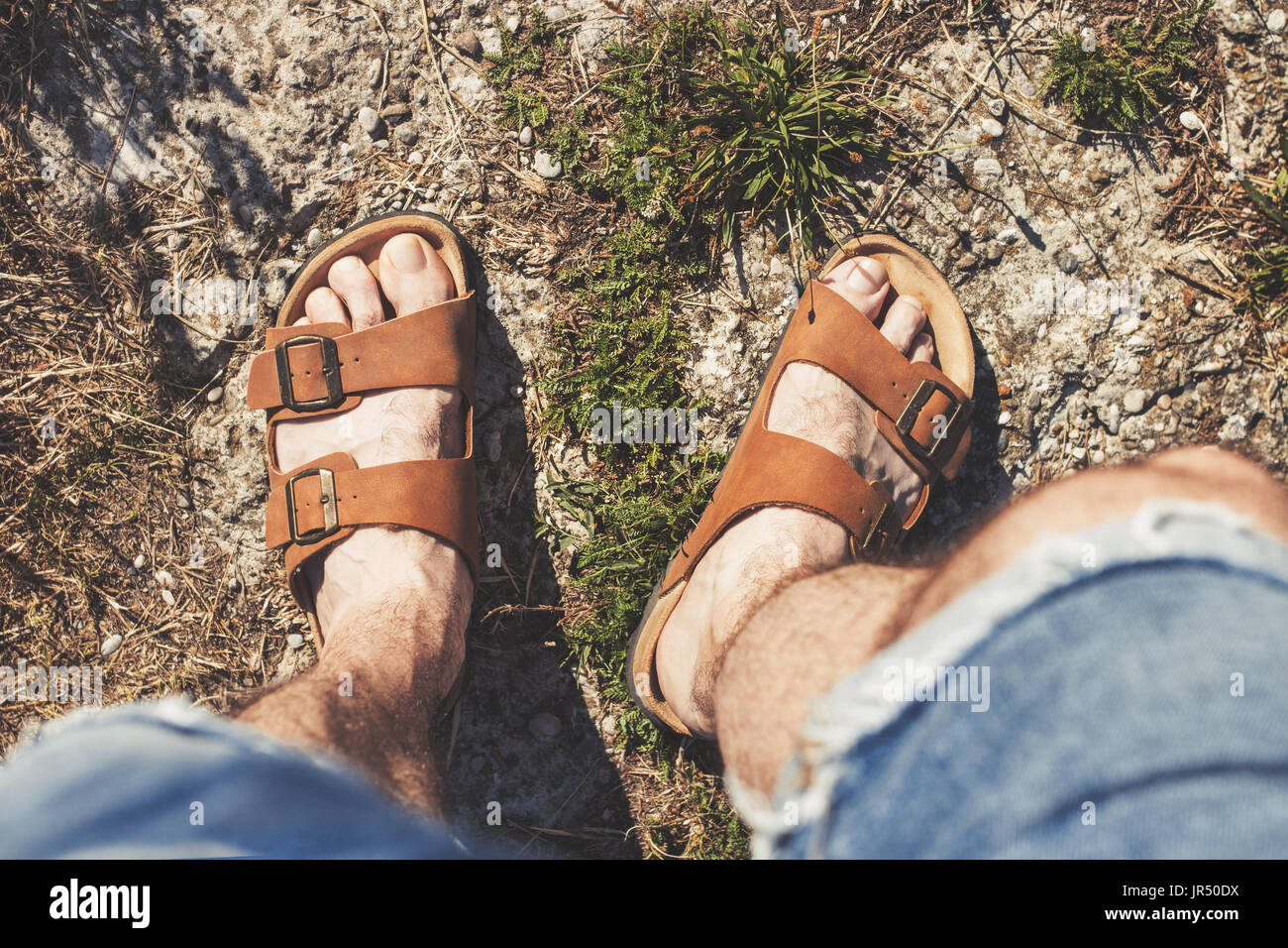 Top view of male legs in brown leather sandals and blue jean shorts, standing on a rocky trail Stock Photo