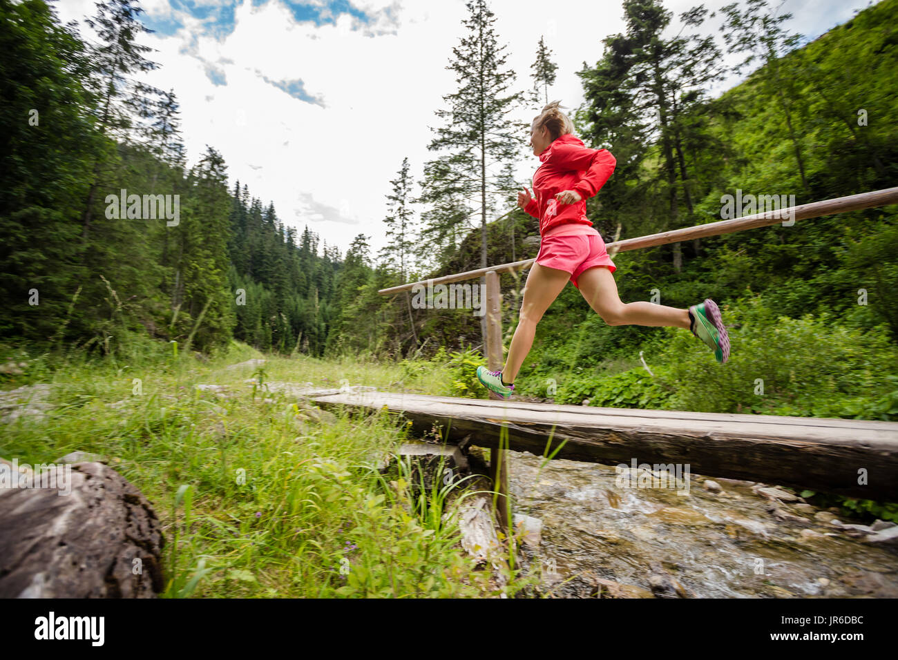 Young woman running in mountains on summer sunny day. Female trail runner crossing bridge on a mountain river. Sport and fitness concept outdoors in n Stock Photo