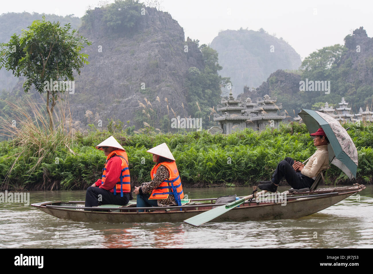 Two tourists are carried on a rowboat by a rower who shelters from the rain under an umbrella in Tam Coc, Vietnam Stock Photo