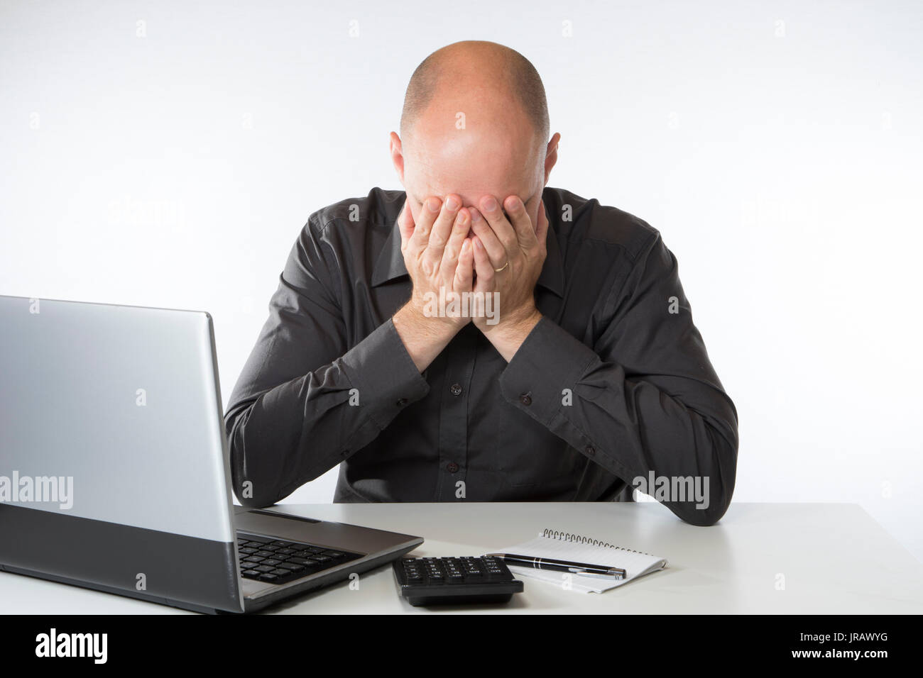 Despairing man doing his accounts sitting at his laptop computer with a calculator and his head in his hands Stock Photo
