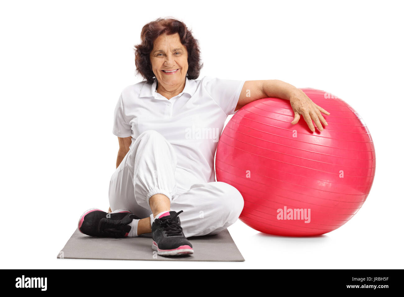 Elderly woman sitting on an exercise mat next to a pilates ball isolated on white background Stock Photo