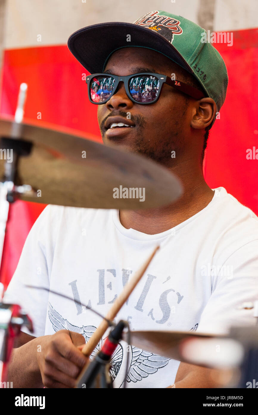 Young afro-caribbean man, 20s, drumming. Wears dark sunglasses and baseball cap, low angle view. Member of group Generation Uncovered. Stock Photo