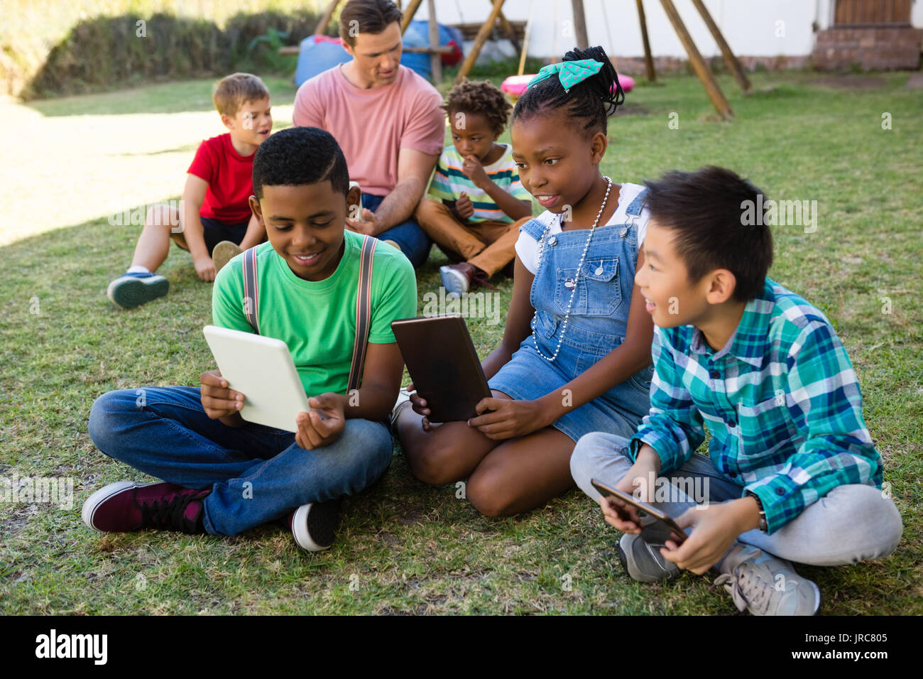 Children using digital tablet while sitting with man on field at park Stock Photo
