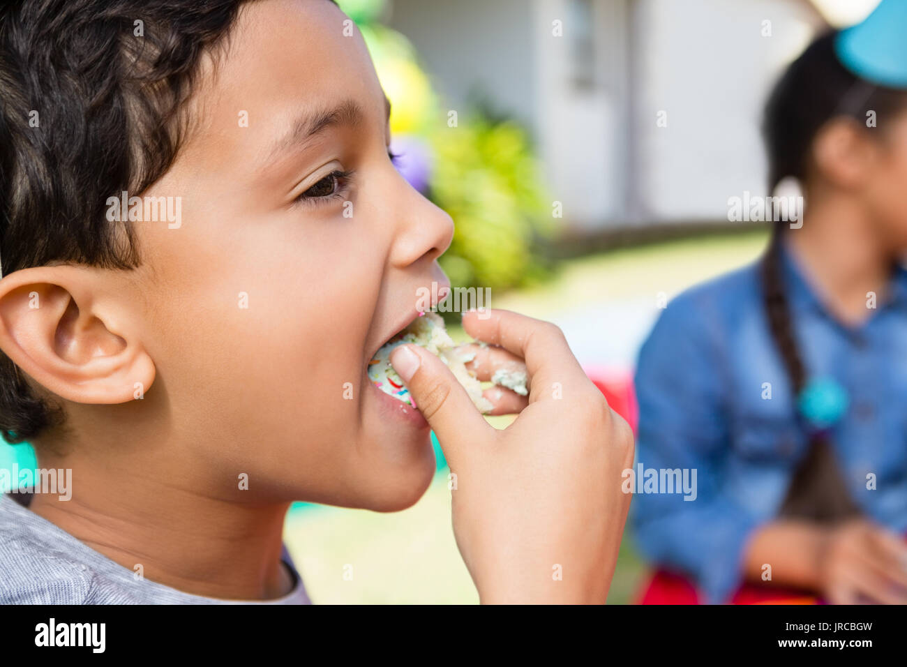 Close up of boy eating cake with friend in background during birthday party Stock Photo