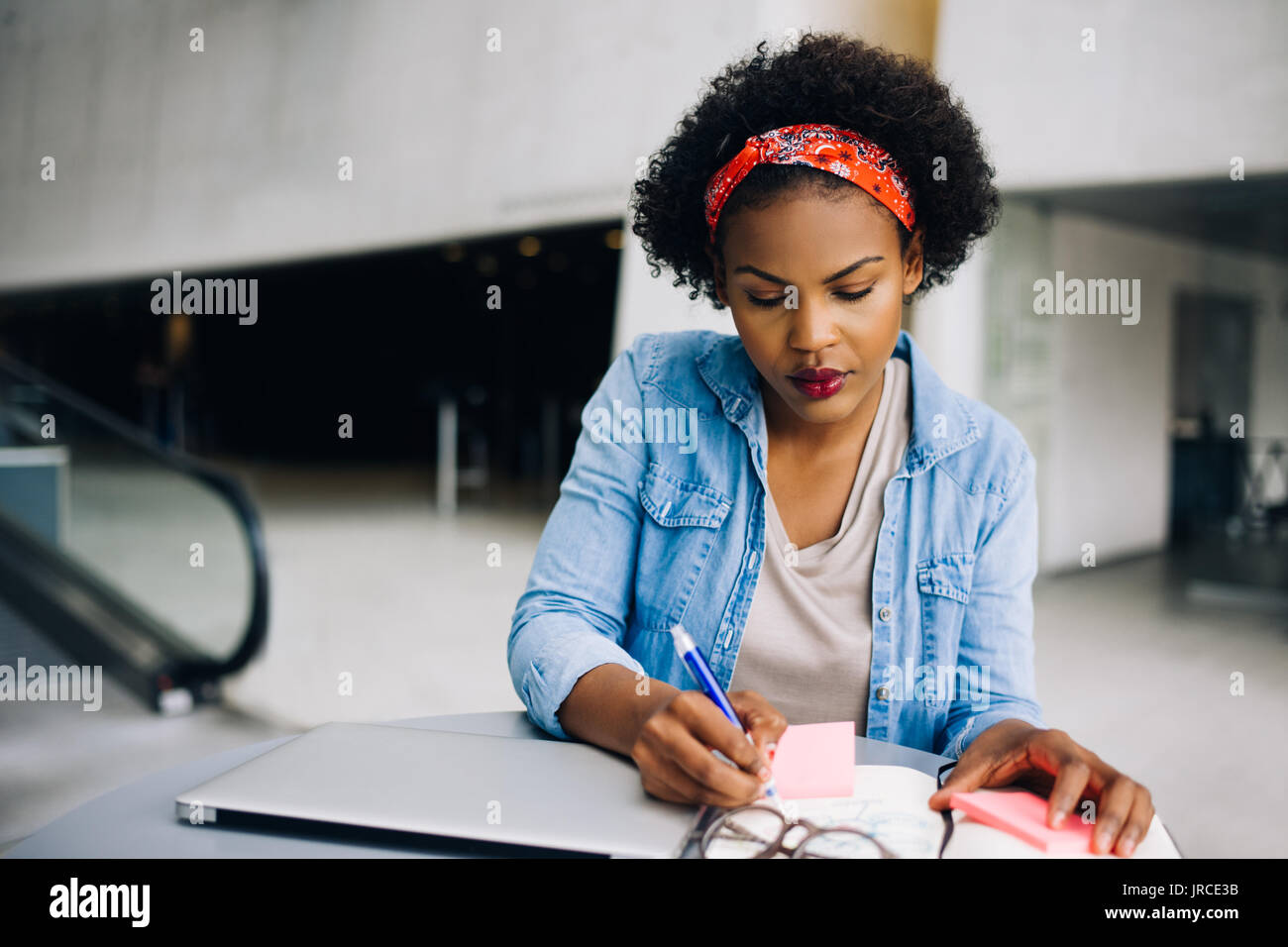 Young African female entrepreneur sitting at a table in a modern office building lobby working on a laptop and writing notes in her planner Stock Photo