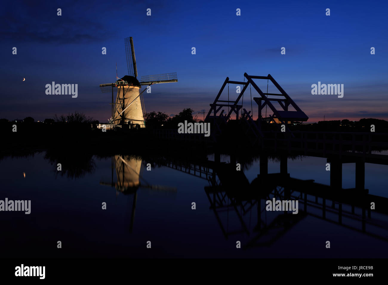 An illuminated windmill and a drawbridge with reflection in the still water of the canal during Blue Hour at sunset in Kinderdijk in the Netherlands. Stock Photo