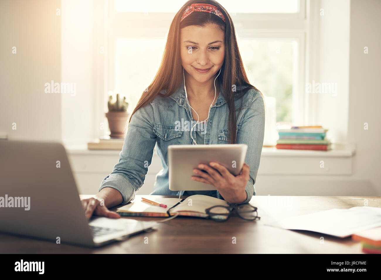 Smiling young entrepreneur woman wearing earphones and browsing tablet while sitting at laptop in office. Stock Photo
