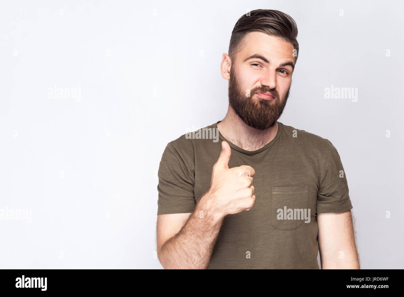 Portrait of satisfied bearded man with thumbs up and dark green t shirt against light gray background. studio shot. Stock Photo