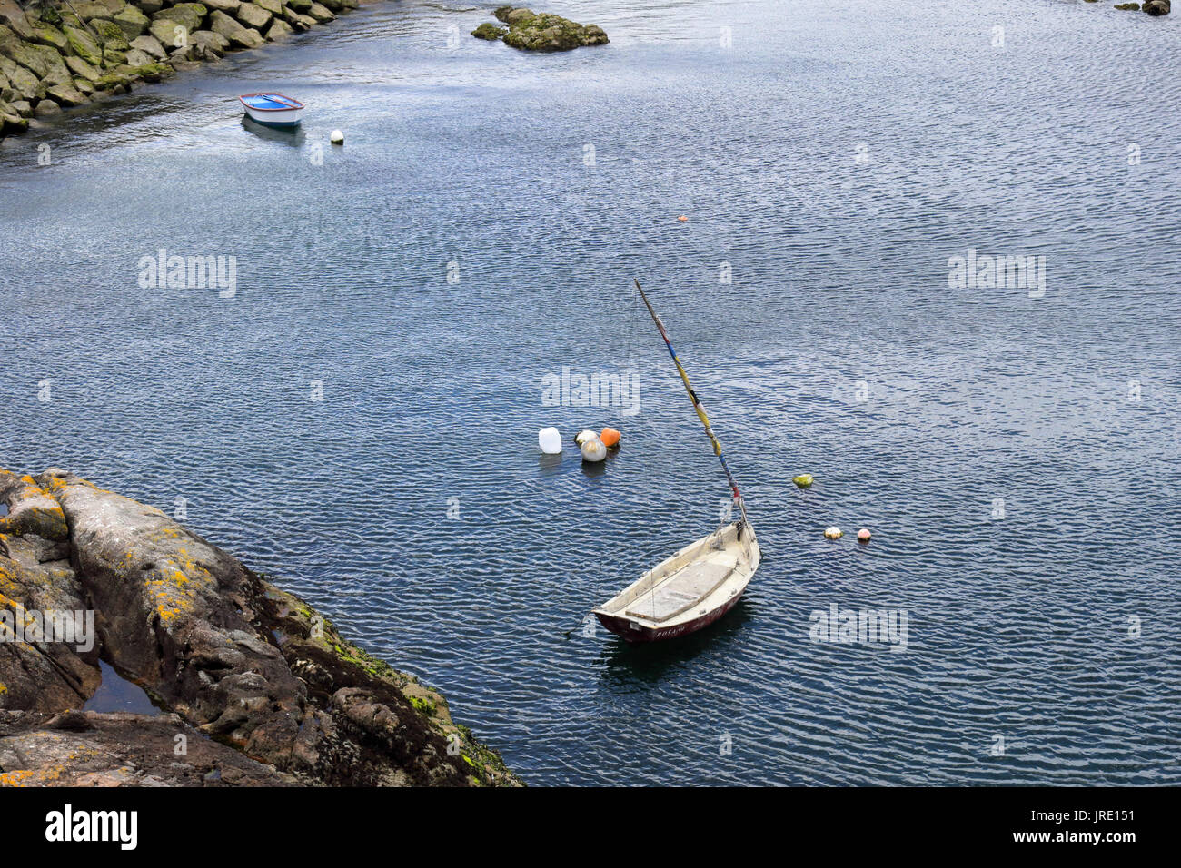 Two small white wooden boats anchored in a port next to a rock pier and cliffs with some buoys. Stock Photo