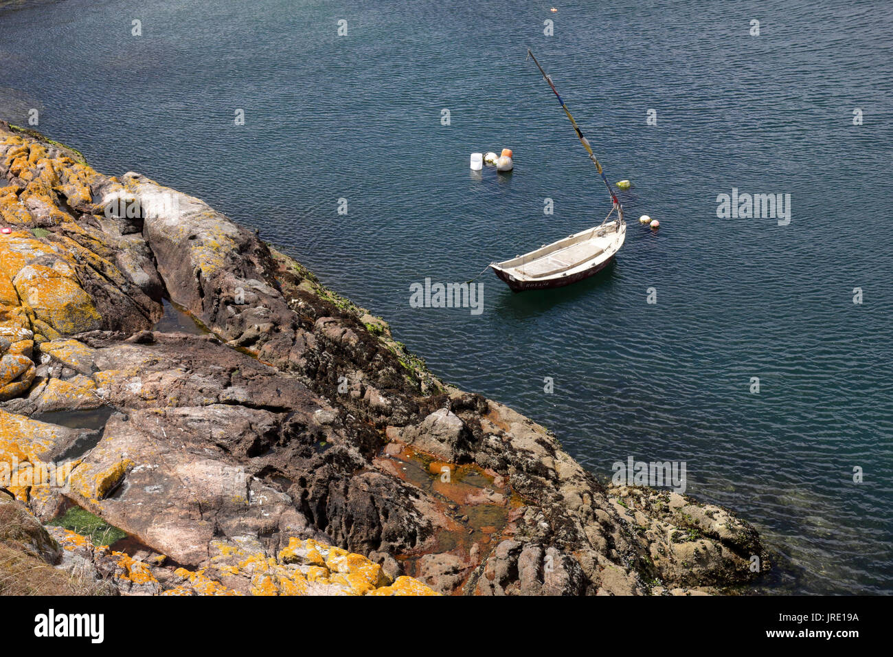 A small white wooden boat anchored in a port next to a rock pier and cliffs with some buoys. Stock Photo
