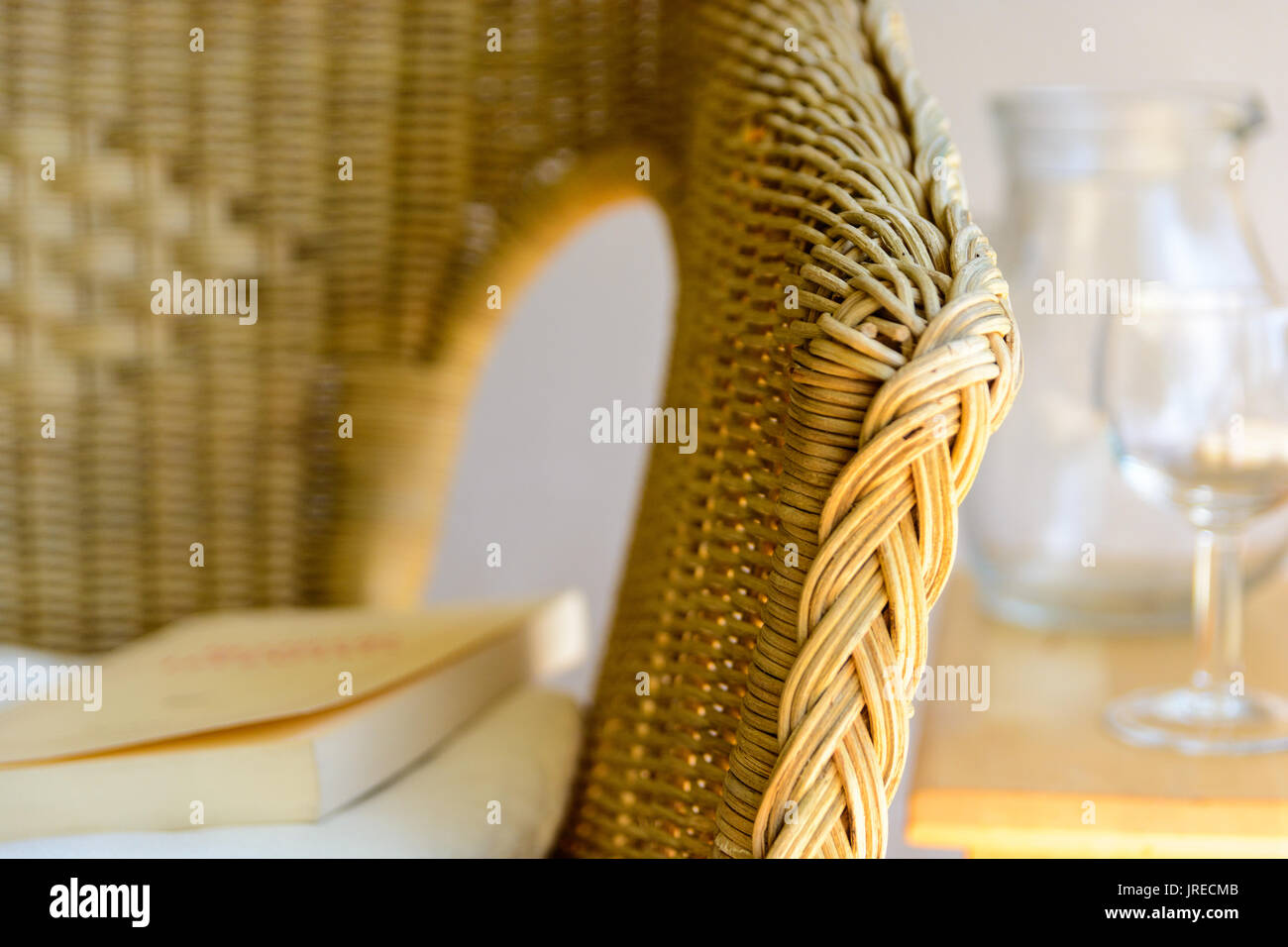 Close-up view on the armrest of a rattan chair with a white cushion, a book, a glass pitcher and a glass of water. Stock Photo