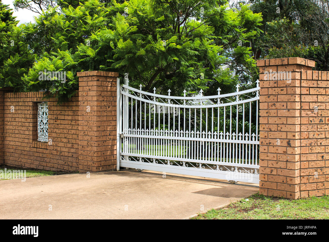 Metal driveway entrance gates set in brick fence with garden trees in background Stock Photo