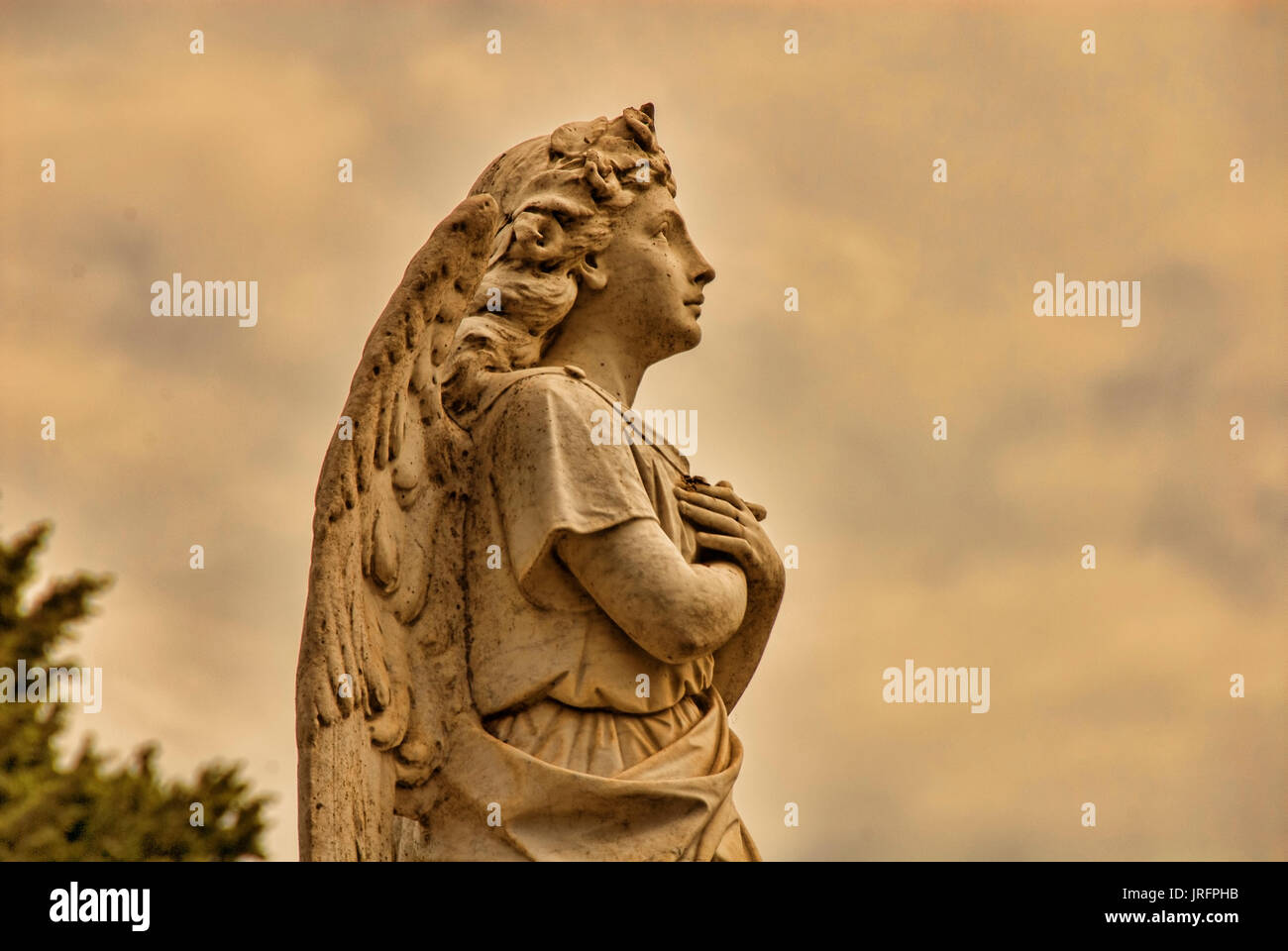 a beautiful stone angel with crossed arms gazes out under a sky full of clouds. Stock Photo