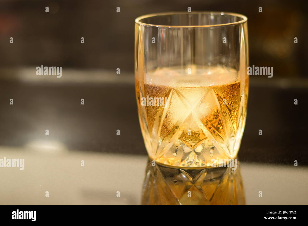 Gold and amber colored Whiskey on the rocks a in a crystal glass, with a giant melting cube of ice on a black granite bar. Stock Photo