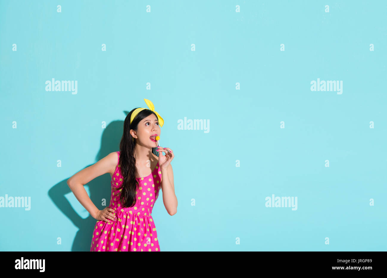 sweet lovely lady eating candy lollipop looking at empty area daydreaming in blue background. Stock Photo