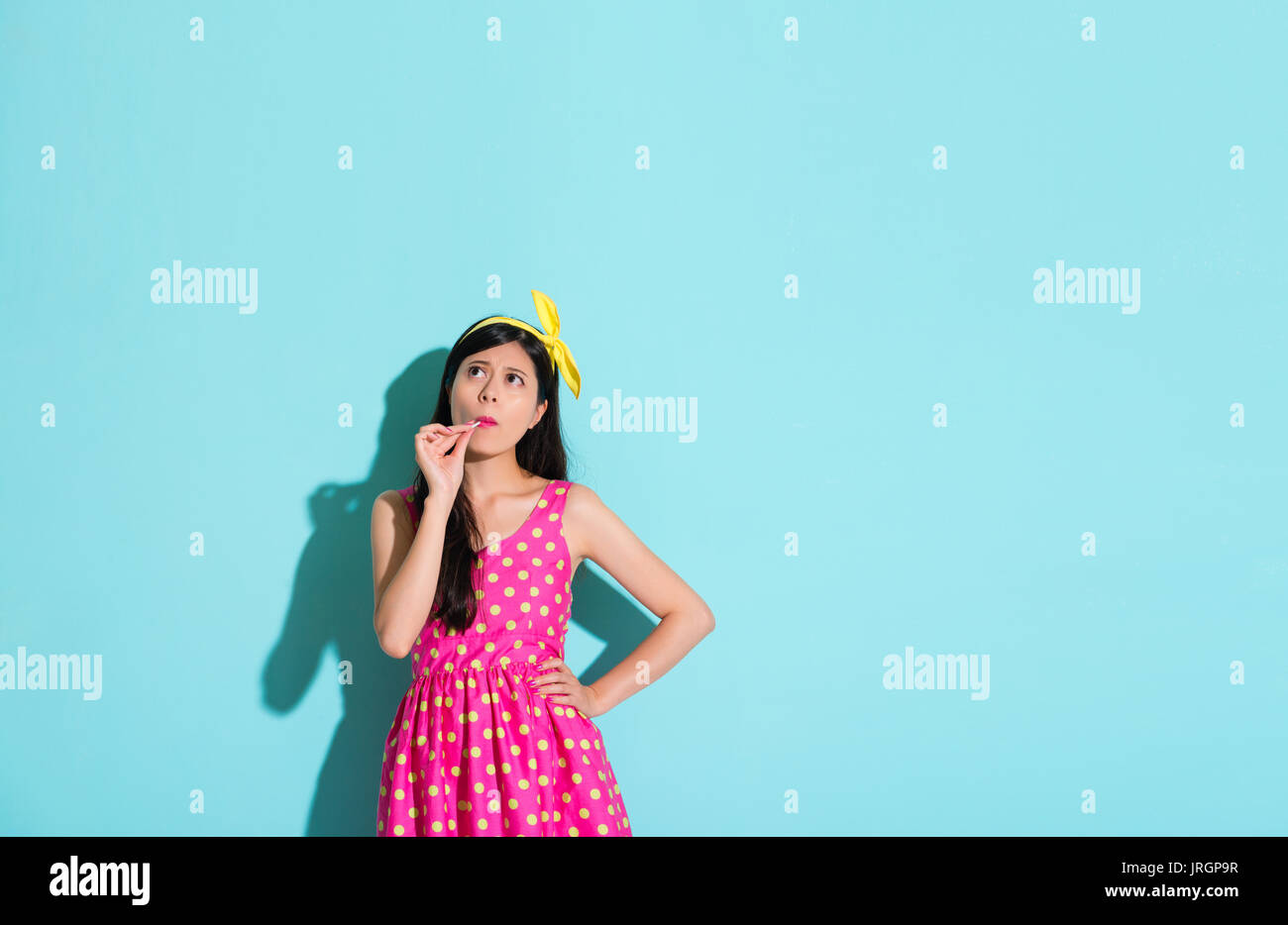 unhappy pretty girl standing in blue background eating lollipop candy and looking at empty area thinking confused problem. Stock Photo