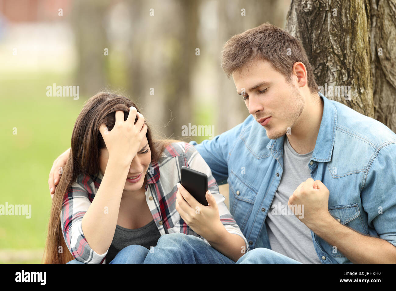 Happy boy celebrating that his sad friend is breaking up by chat sitting on the grass in a park Stock Photo