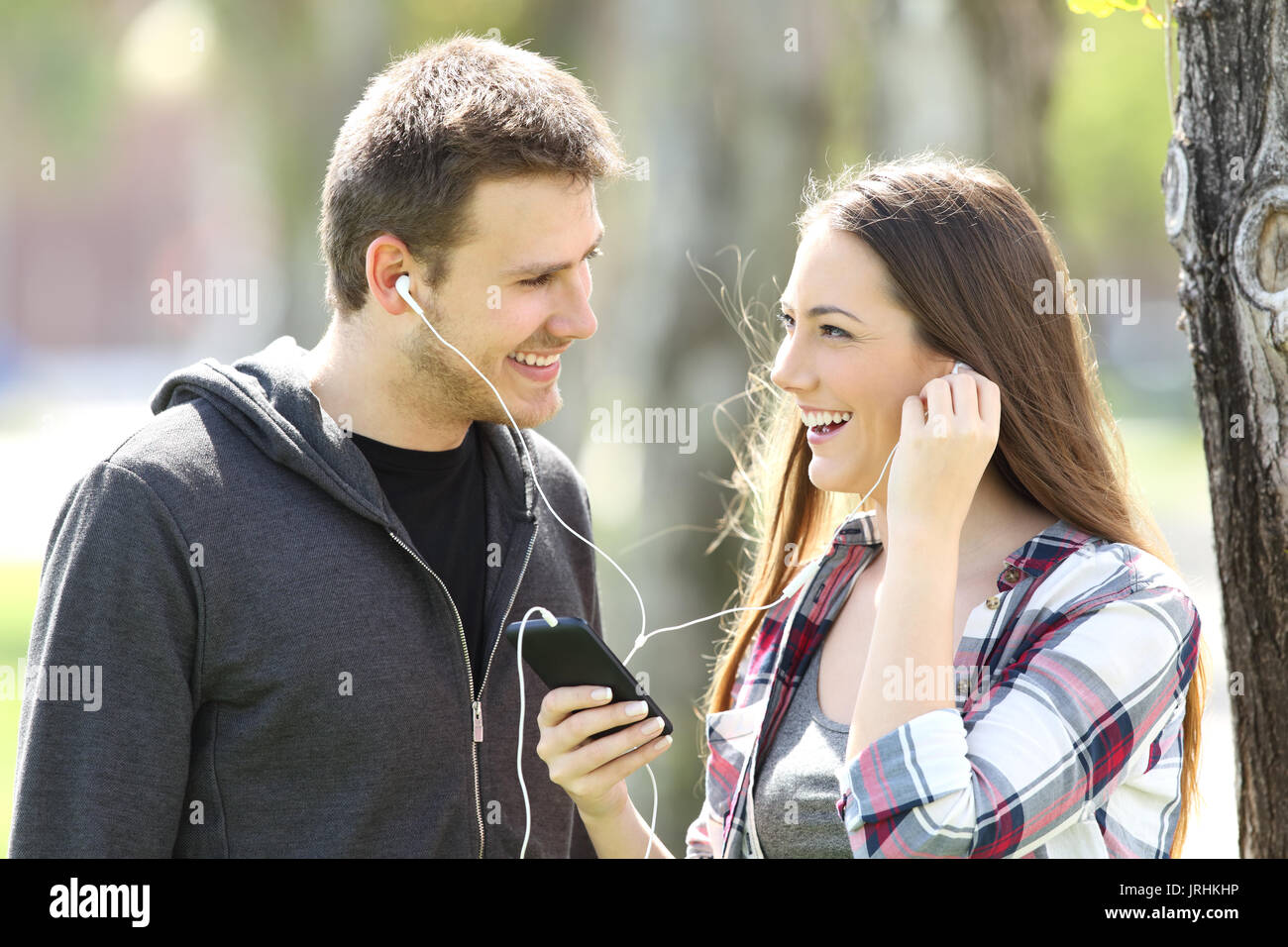 Happy couple of teens sharing on line music in a smart phone outdoors in a park Stock Photo