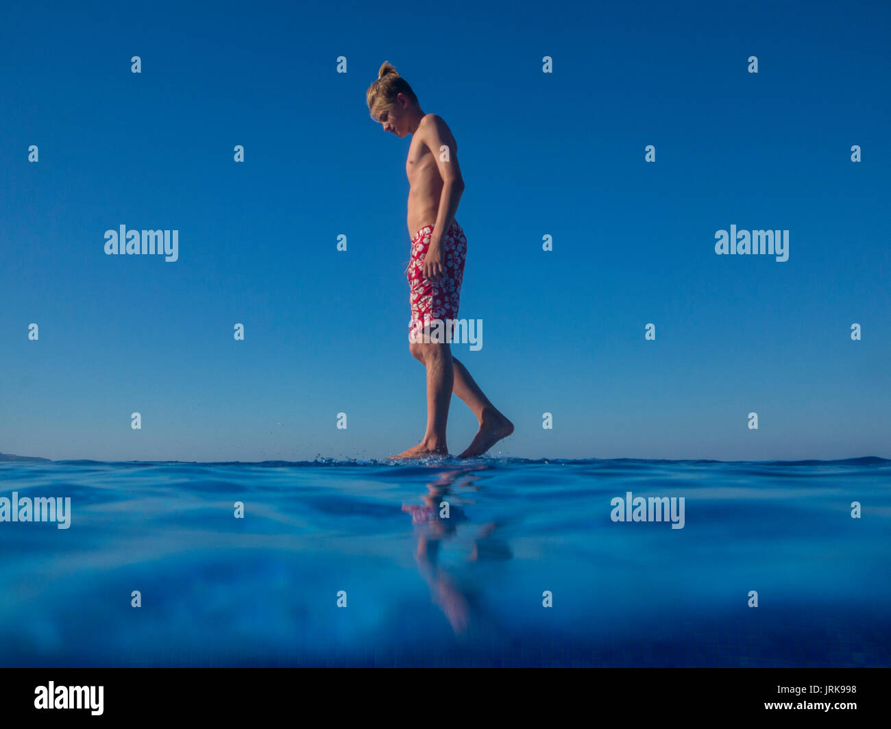 A bored teenaged boy walks on the edge of an infinity pool on holiday Stock Photo