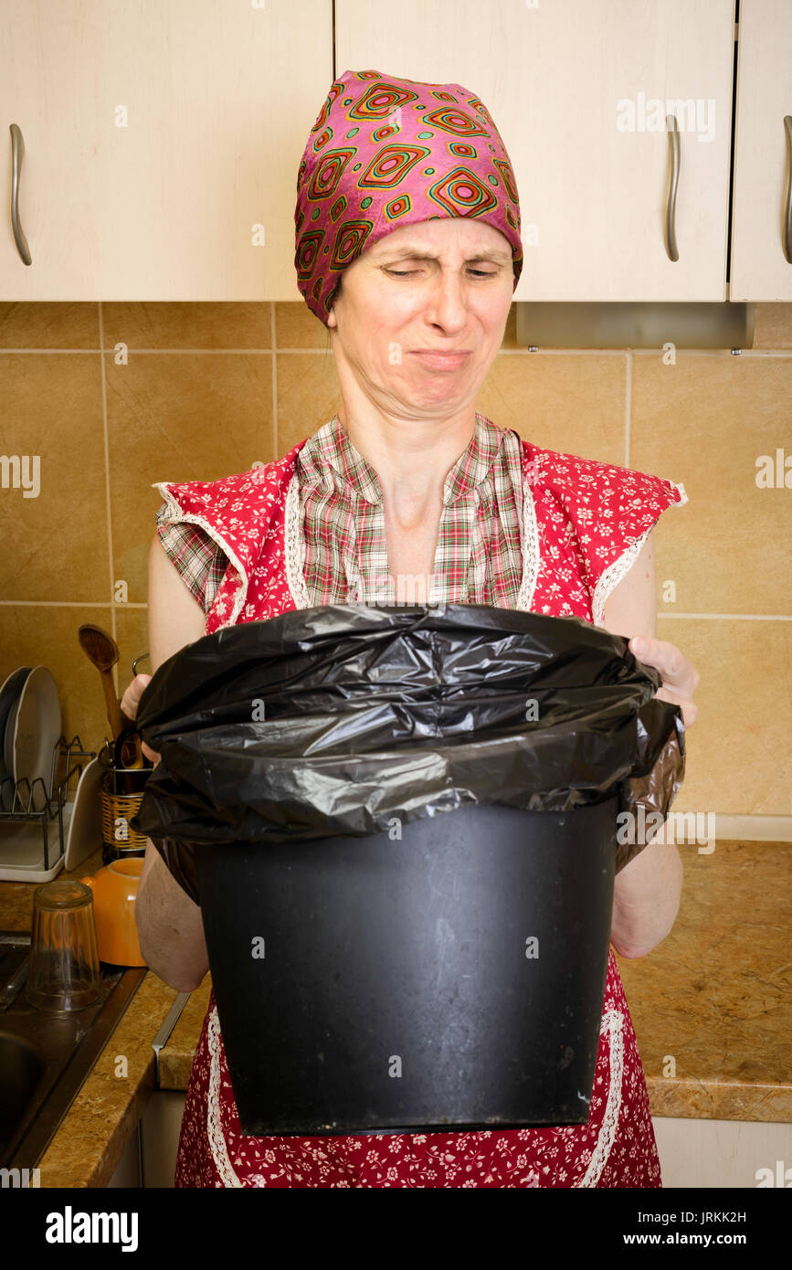 A woman, with a scarf on the head and a red apron, is looking inside a black trash can with a garbage bag, in the kitchen. She is very disturbed by th Stock Photo