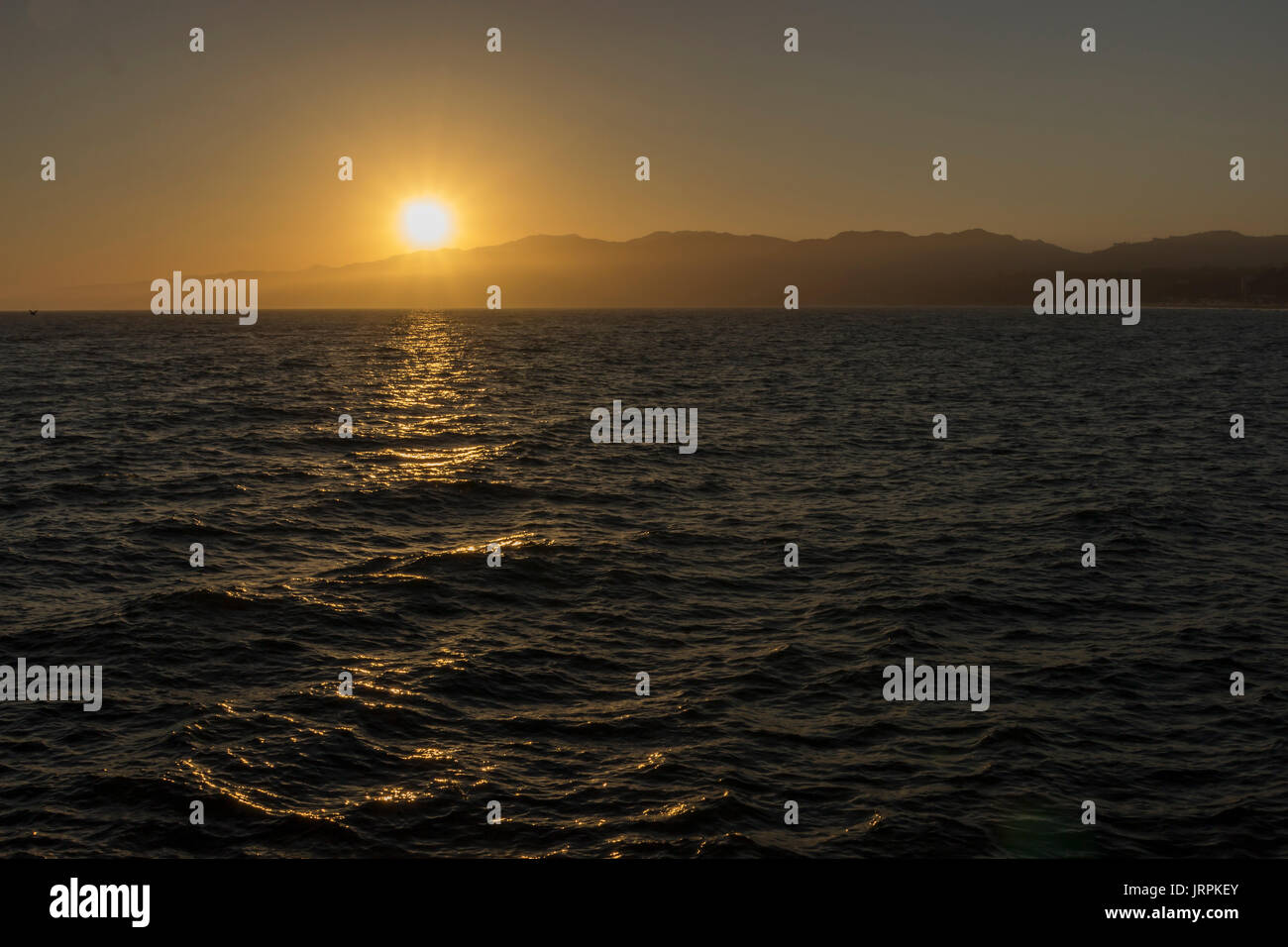 Anochecer en el oceano pacifico, en las pliayas de Santa Monica, el reflejo del sol en el agua, deja un camino de luz, atras se ven pequeñas  montañas Stock Photo