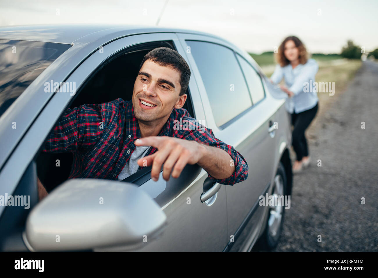 Woman pushing a broken car down the road, smiling man driver. Vehicle with trouble Stock Photo