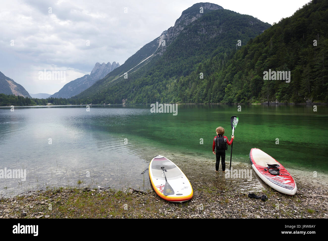 Young boy with dark blonde hair standing in the water between SUP, holding a paddle and looking to the mountains at lake Lago di Predil, Italy. Stock Photo