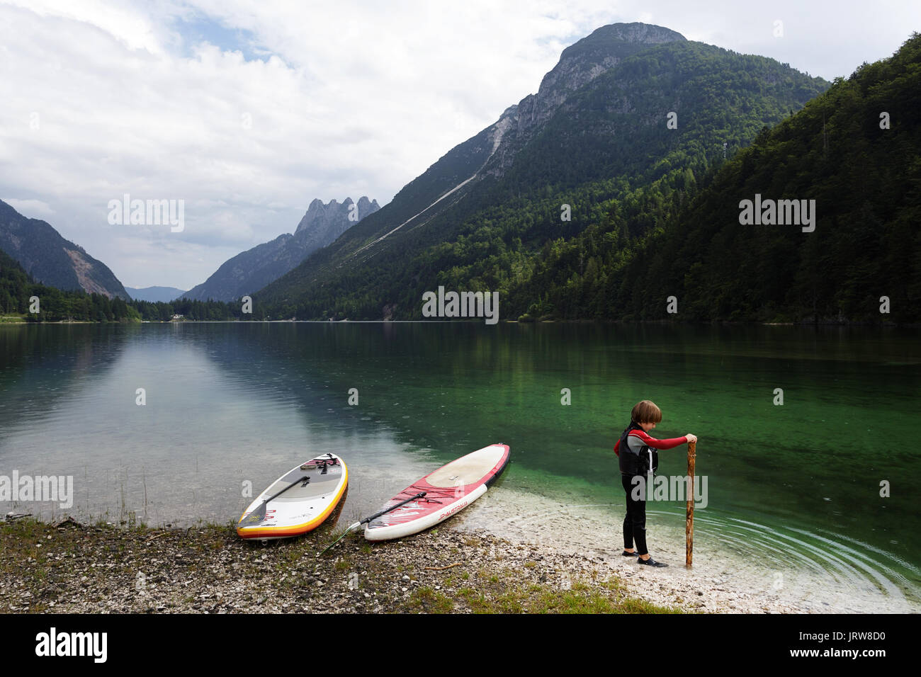 Young boy in a neoprene suit and life jacket playing with a large stick by SUP at alpine lake Lago di Predil, Italy. Stock Photo