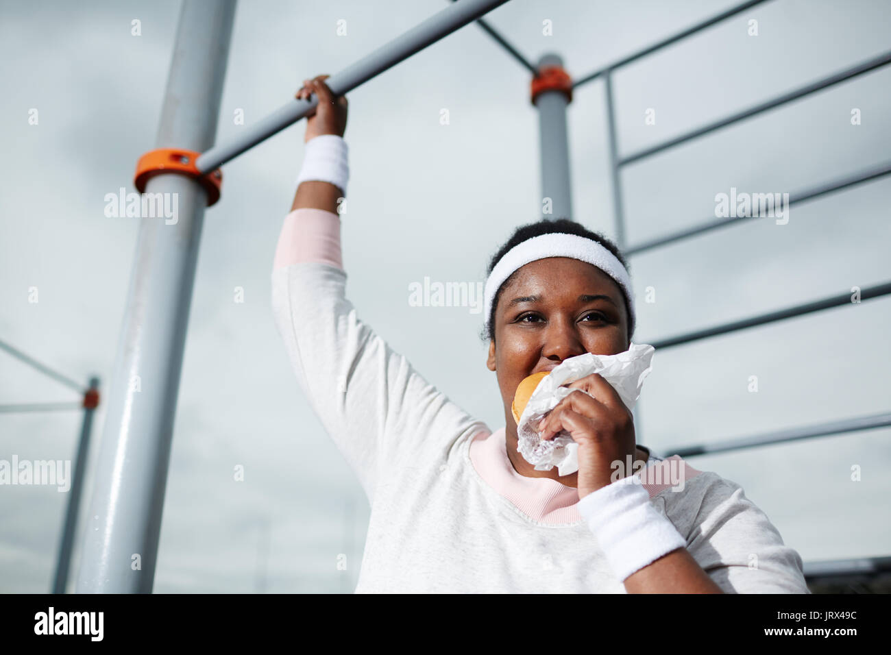 Plump African woman eating unhealthy burger while practicing pull-up exercise Stock Photo