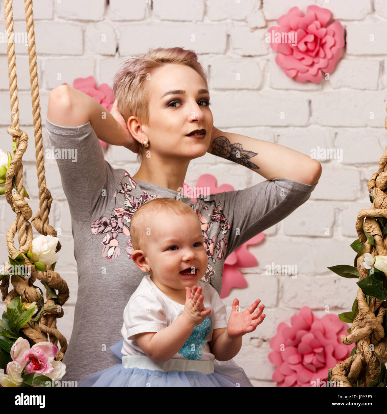 Attractive young mother with her small daughter on swing on light brick`s background Stock Photo