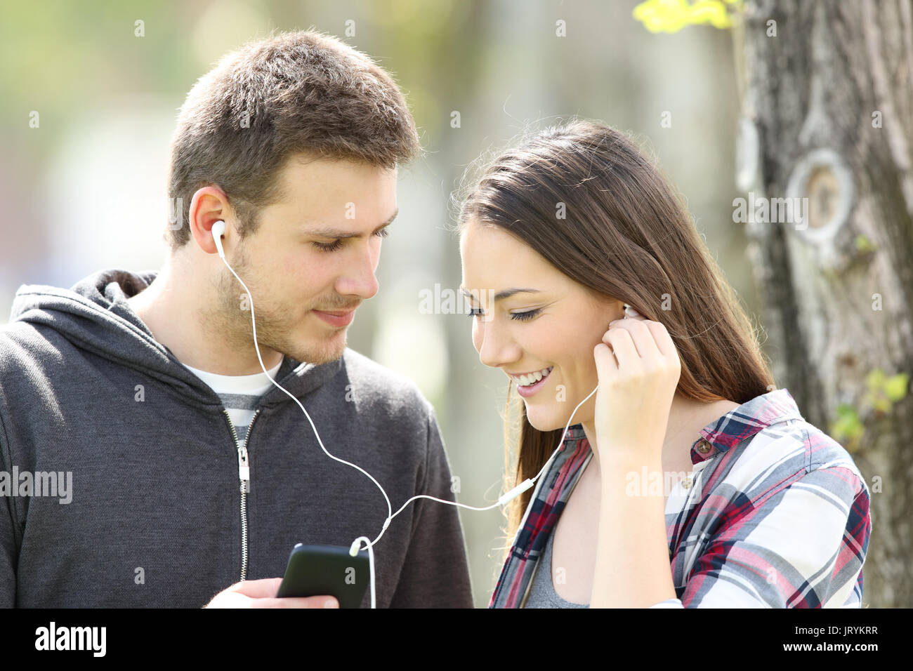 Couple flirting and sharing music on line with earphones and a smart phone outdoors in a park Stock Photo
