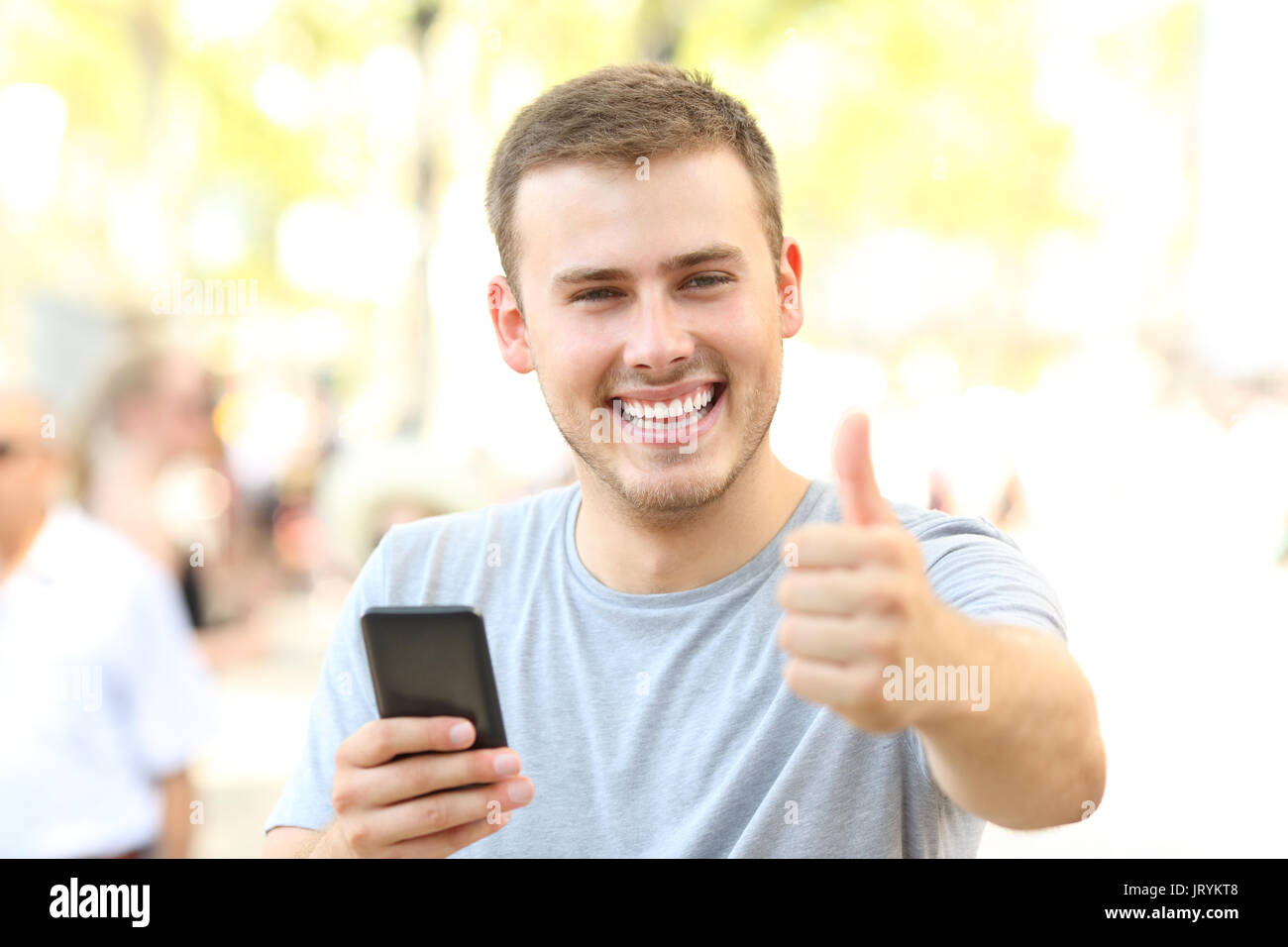 Man with thumbs up holding phone looking at you on the street Stock Photo