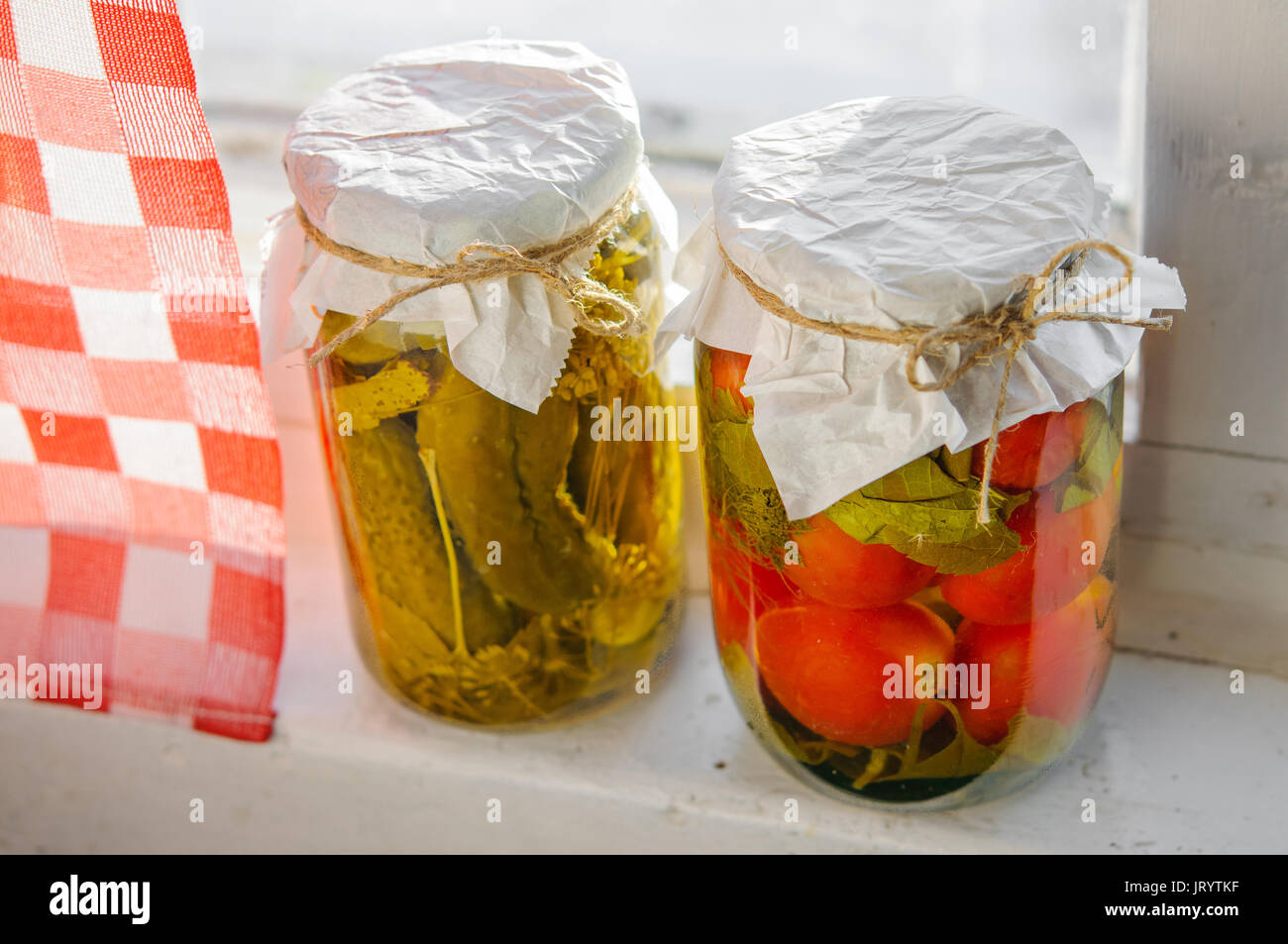 Clear glass jars of pickled vegetables at window sill: tomatoes and cucumbers in rustic style Stock Photo