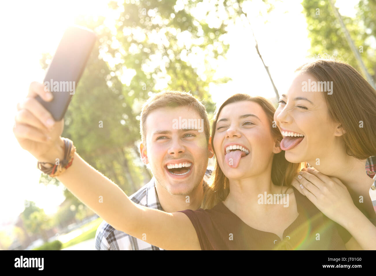 Back light of three funny teens taking selfies and joking in the street Stock Photo