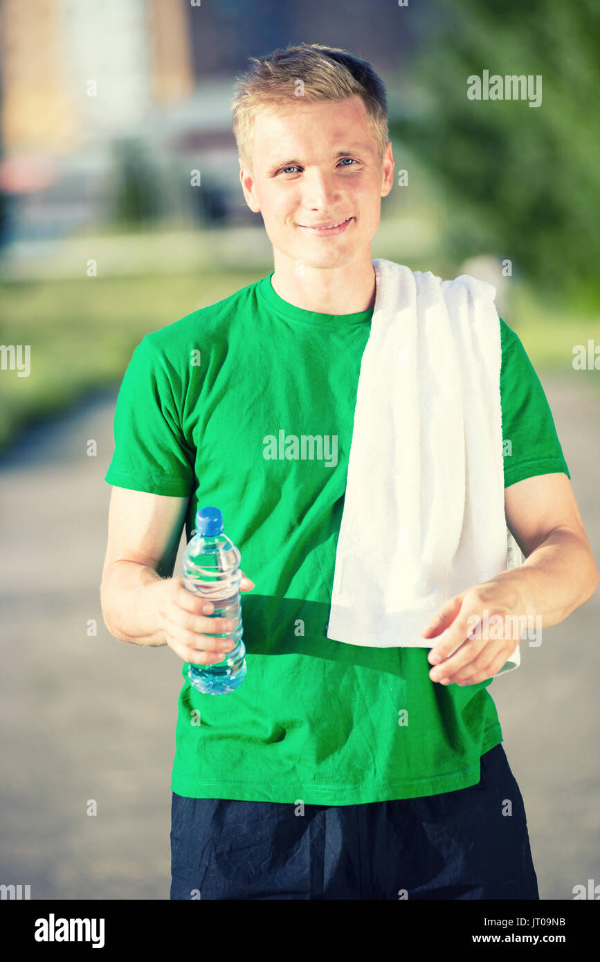 Tired man with white towel drinking water from a plastic bottle after fitness time and exercising in city street park at beautiful summer day. Sporty  Stock Photo