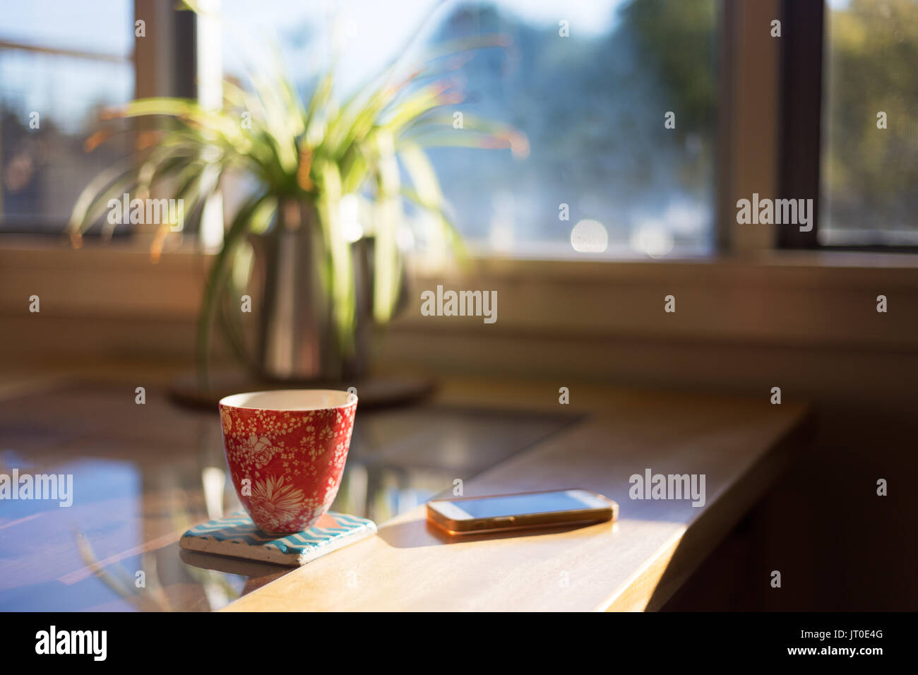 A cup of coffee on a coaster next to smart phone on table with bright morning window light streaming through. Stock Photo