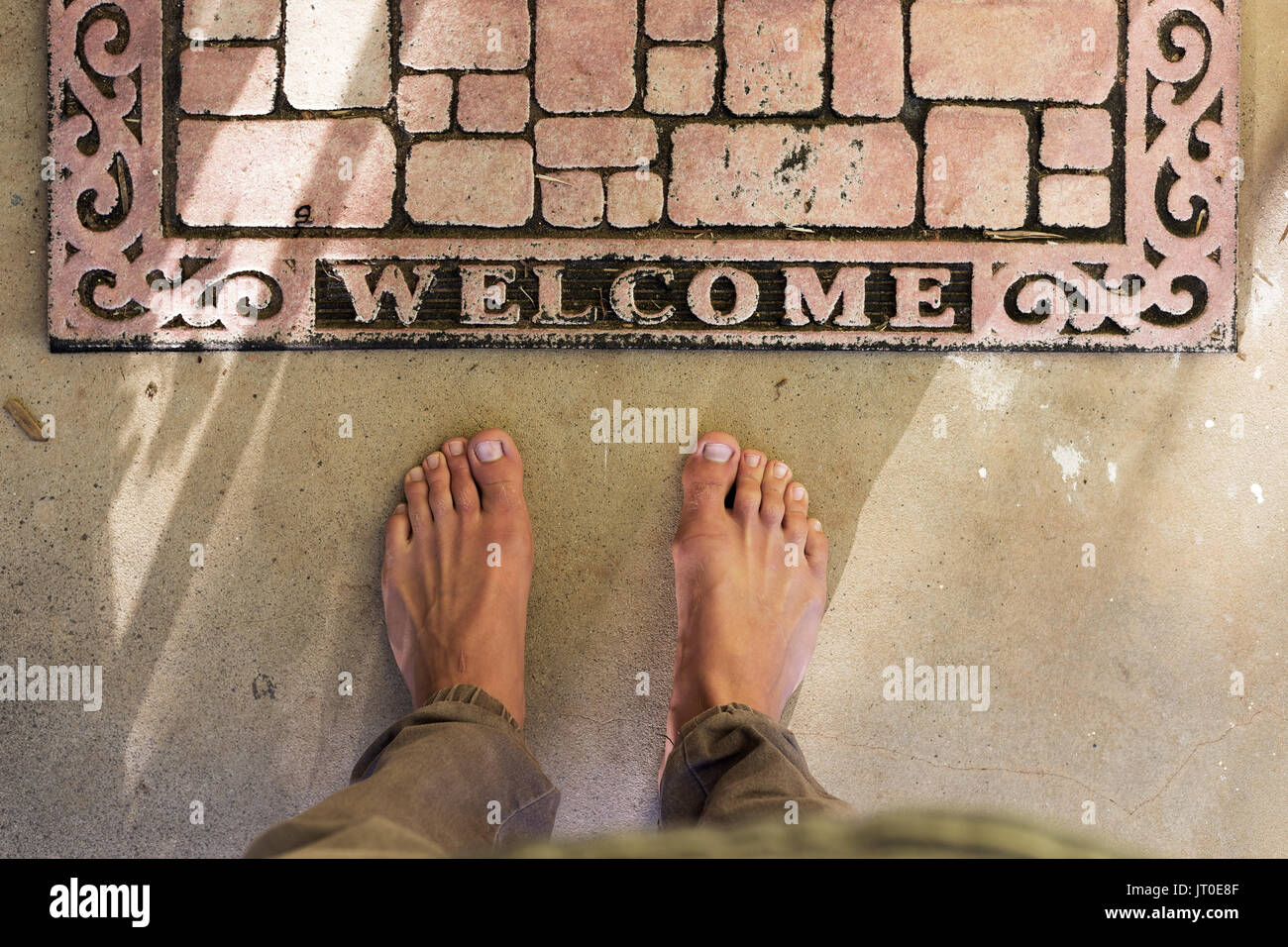 Point of view of bare feet at home front door welcome mat on concrete patio. Stock Photo