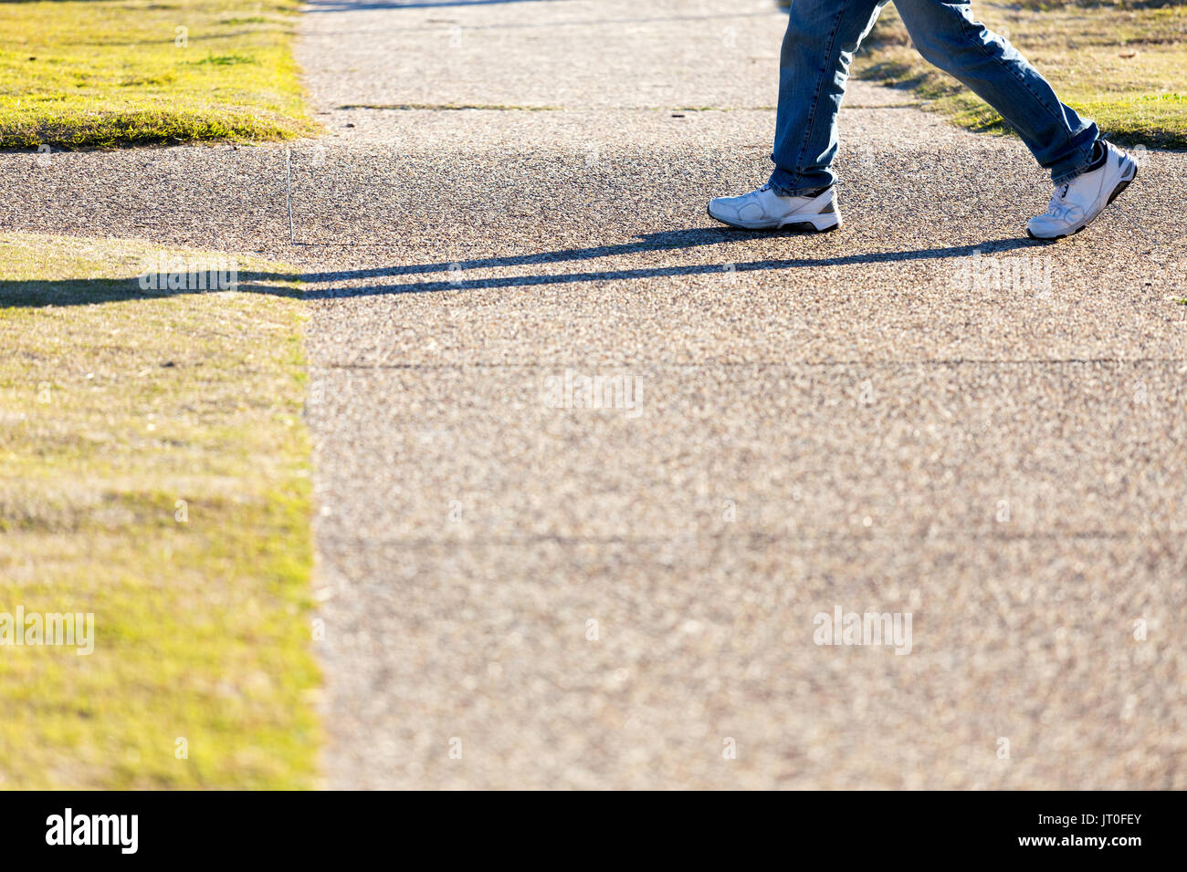 A below waist shot of a man wearing blue jeans and white sneakers walking past a footpath intersection in a city park. Stock Photo