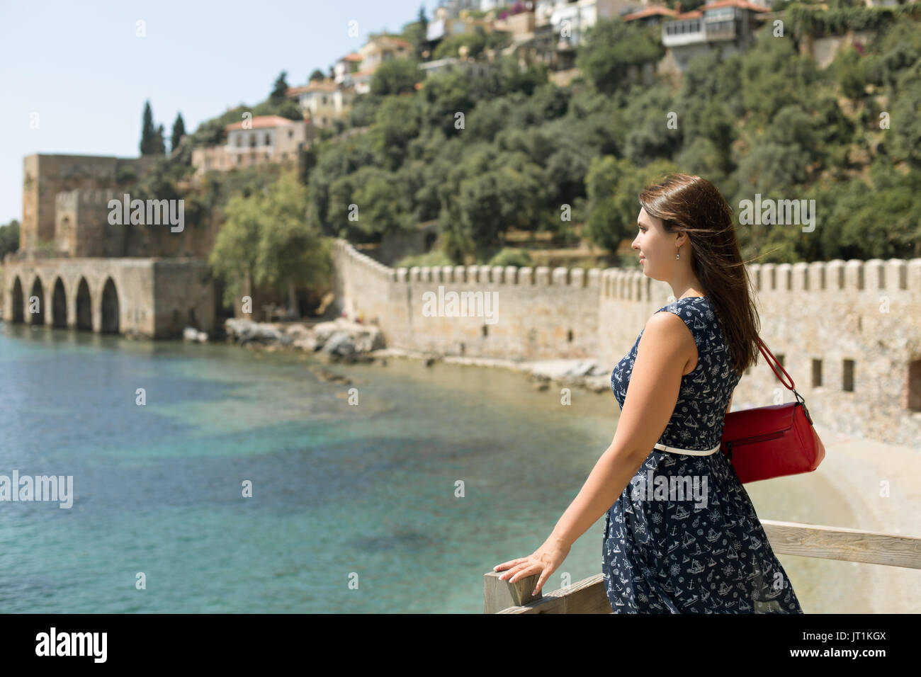 SIdeview portrait of beautiful woman with strait hair wearing dark blue dress  with white belt and red bag enjoying view of picturesque bay of Alanya  Stock Photo
