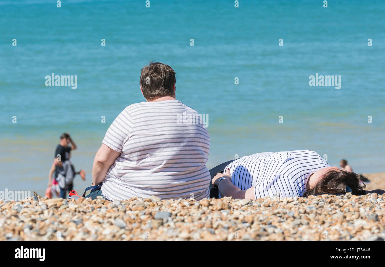 Overweight woman sitting on a beach. Large lady sat in the sun on a beach in the UK. Obese person on a beach. Stock Photo