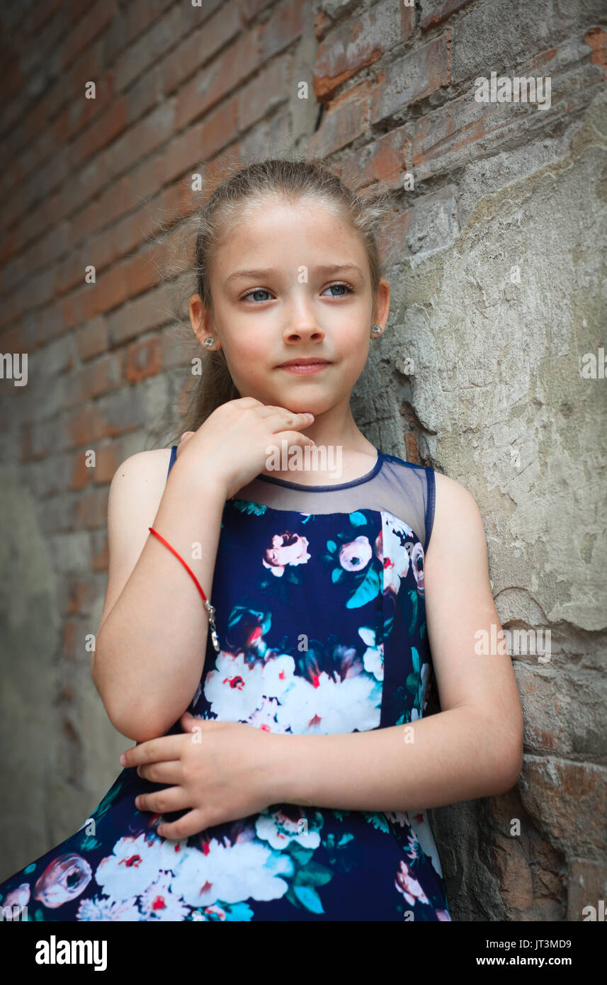 Portrait of a cute young school-age girl in a beautiful dress outdoors against a brick wall background. Stock Photo