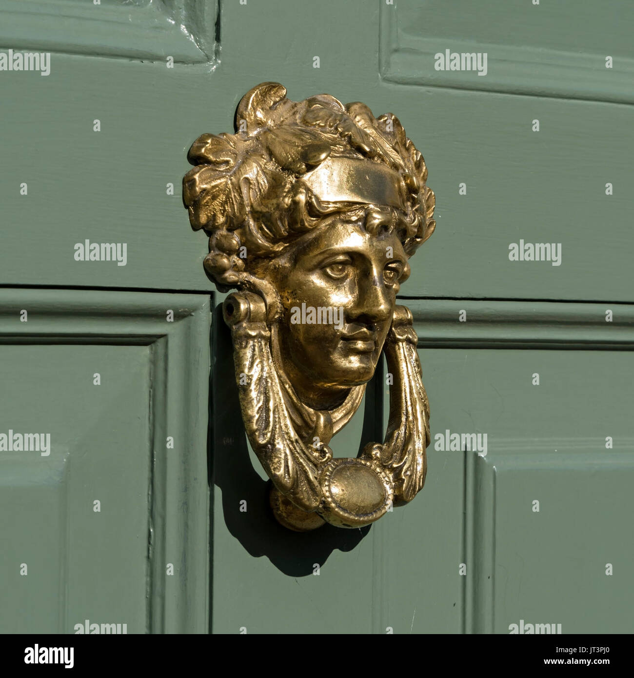 Smart golden polished brass door knocker depicting Roman goddess Apollo, on green painted wooden panelled front door. Stock Photo