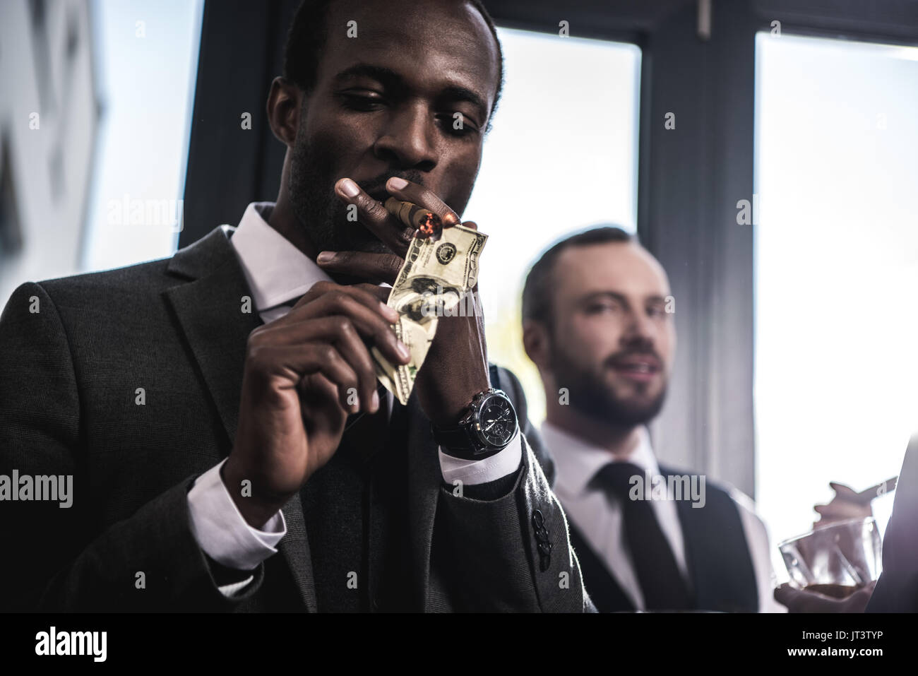 Confident businessman lighting cigar with dollar banknote indoors Stock