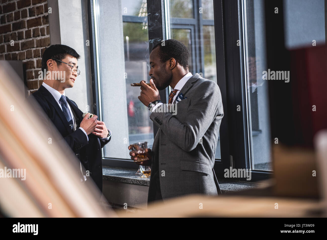 Confident businessman drinking alcohol beverage and smoking cigar while colleague hiding money into suit pocket, business team meeting Stock Photo