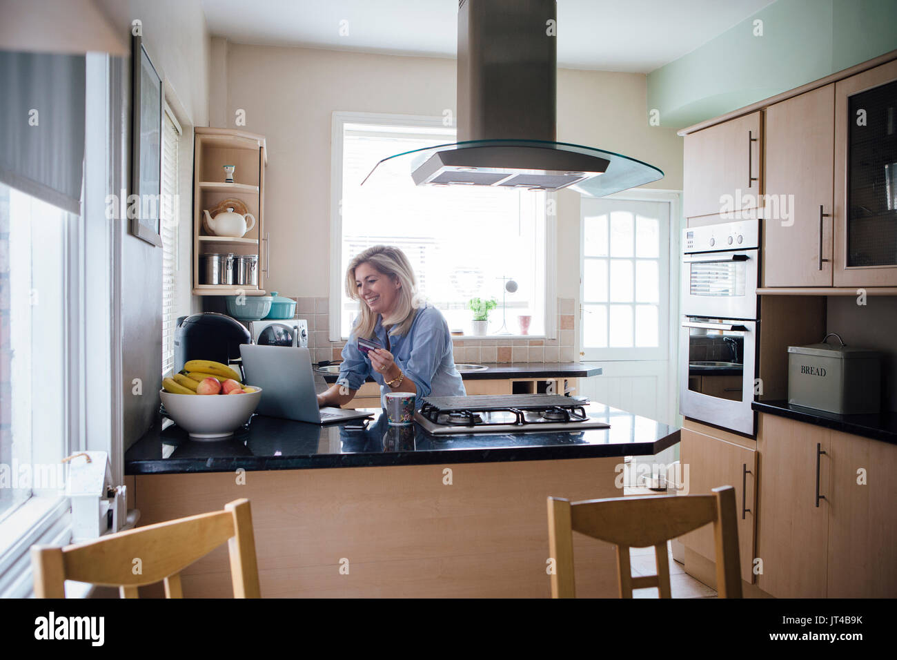 Mature woman using her laptop in her kitchen to make an online payment with a credit card. Stock Photo